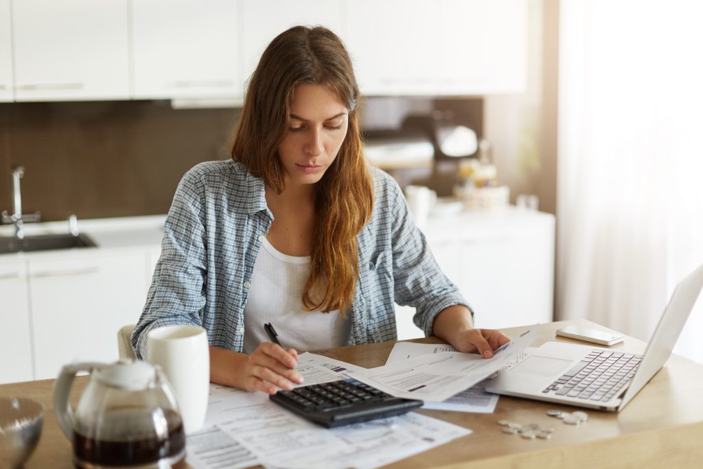 Jeune femme concentrée faisant ses comptes à la table de sa cuisine, avant de faire une demande de prêt rapide, utilisant une calculatrice et un ordinateur portable, entourée de documents et de reçus.