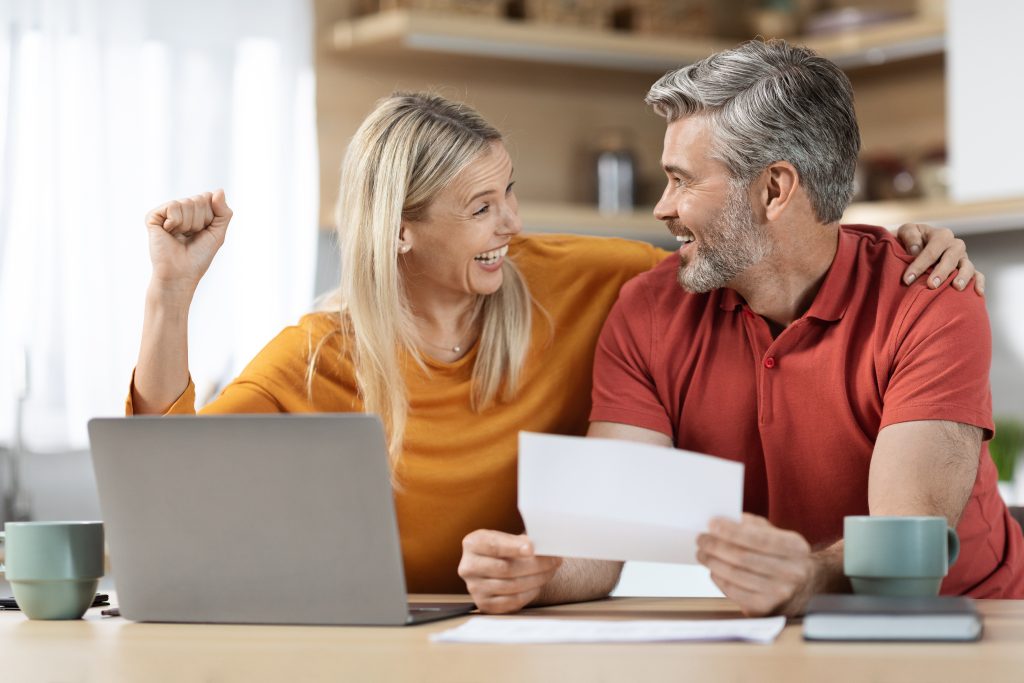 Comment trouver de l'argent quand les banques refusent? - dmocredit Couple souriant à la maison, célébrant une bonne nouvelle financière après avoir obtenu un prêt rapide sans documents en ligne devant un ordinateur portable. Comment trouver de l'argent quand les banques refusent.