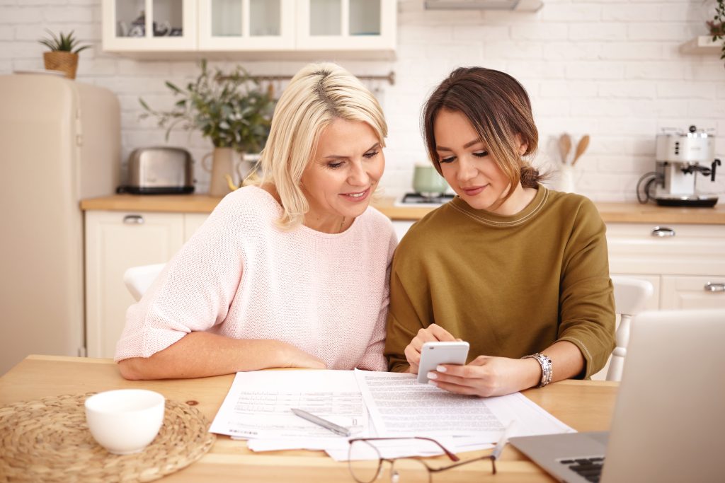 Two women sitting at a kitchen table, reviewing financial documents and using a smartphone, discussing the responsible use of a payday loan.