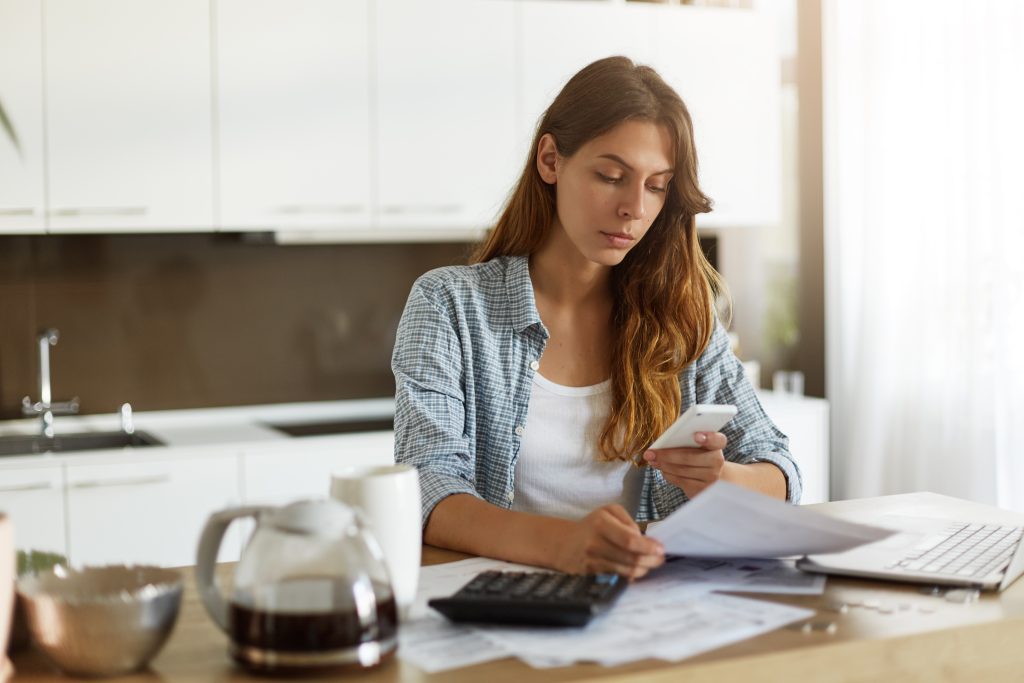 Young woman at home reviewing her finances with a calculator, documents, and her phone, looking for a $500 loan to manage an urgent expense.